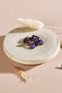 Boutons de forme irrégulière façon pierre facettée, aspect satiné, coloris violet profond, avec fixation à queue métallique dorée, présentés en tas sur une petite coupelle en céramique blanche, sur fond rose avec des épis de lagurus séchés.