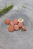 Boutons de forme irrégulière façon pierre facettée, aspect nacré, coloris rose pêche melba, avec fixation à queue métallique dorée, posés sur une pierre grise avec un brin de romarin en arrière-plan.