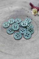 A dozen small four-hole round buttons in glossy opaque cactus green, stacked on grey stone with dried flowers in the upper right.