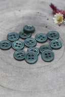Douzaine de petits boutons ronds à quatre trous vert cèdre grisé brillant, empilés sur pierre grise avec fleurs séchées.