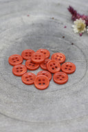 A dozen small four-hole round buttons in vivid glossy orange (poppy tangerine), stacked on grey stone with dried flowers in the background.