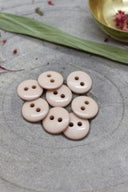 Classic two-hole round buttons, glossy taupe-pink blush finish, piled on grey stone, with a green leaf and dried petals in the background.