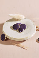 Four domed round and oval buttons, glossy finish, in deep violet, rimmed with a thin gold trim, displayed in a small white ceramic dish on a pink background with a dried lagurus spike.