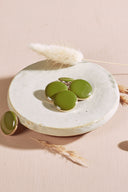 Four domed round and oval buttons, glossy finish, in matcha green, rimmed with a thin gold trim, displayed in a small white ceramic dish on a pink background with a dried lagurus spike.