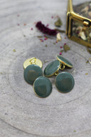 Cinq boutons ronds facettés vert cèdre (vert sauge foncé) cerclés d'un liseré doré, posés sur une pierre grise avec fleurs séchées et un coffret doré flou en arrière-plan.