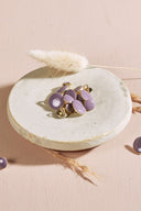 Boutons ronds bombés mauve parme cerclés d'un fin liseré doré, à queue métallique (shank), présentés en tas dans une petite coupelle en céramique blanche sur fond rose, avec un brin d'herbe séchée (lagurus) en arrière-plan.