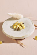 Domed round buttons in lemon yellow rimmed with a thin gold trim, with a metal shank, piled in a small white ceramic dish on a pink background, with a sprig of dried lagurus grass in the background.