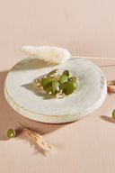Boutons ronds bombés vert matcha cerclés d'un fin liseré doré, à queue métallique (shank), présentés en tas dans une petite coupelle en céramique blanche sur fond rose, avec un brin d'herbe séchée (lagurus) en arrière-plan.