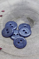 Boutons ronds à deux trous entièrement pailletés bleu nuit, présentés en tas sur une pierre grise, avec un fil de raphia bordeaux en arrière-plan.