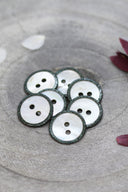Boutons ronds à deux trous en nacre blanche, cerclés d'un liseré pailleté vert sombre, présentés en tas dans une coupelle en pierre grise, avec des pétales de fleurs blanches et bordeaux en arrière-plan.