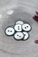 Boutons ronds à deux trous en nacre blanche, cerclés d'un liseré pailleté anthracite, présentés en tas dans une coupelle en pierre grise, avec des pétales de fleurs blanches et bordeaux en arrière-plan.