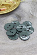 Boutons ronds à deux trous en émail brillant vert cèdre, présentés en tas sur une pierre grise, avec une coupelle dorée contenant des fleurs séchées et un verre facetté en arrière-plan.