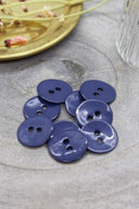 Boutons ronds à deux trous en émail brillant bleu cobalt, présentés en tas sur une pierre grise, avec une coupelle dorée contenant des fleurs séchées et un verre facetté en arrière-plan.