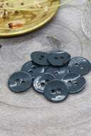 Boutons ronds à deux trous en émail brillant bleu-gris forêt sombre, présentés en tas sur une pierre grise, avec une coupelle dorée contenant des fleurs séchées et un verre facetté en arrière-plan.