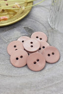 Two-hole round buttons in glossy sandy beige-pink enamel, piled on grey stone, with a gold dish holding dried flowers and a faceted glass in the background.