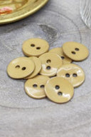 Boutons ronds à deux trous en émail brillant moutarde, présentés en tas sur une pierre grise, avec une coupelle dorée contenant des fleurs séchées et un verre facetté en arrière-plan.