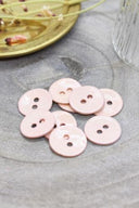Boutons ronds à deux trous en émail brillant rose poudré très pâle, présentés en tas sur une pierre grise, avec une coupelle dorée contenant des fleurs séchées et un verre facetté en arrière-plan.