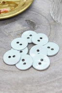Two-hole round buttons in glossy very pale glacier blue enamel, piled on grey stone, with a gold dish holding dried flowers and a faceted glass in the background.