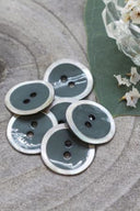 Boutons ronds à deux trous, finition émaillée gris cèdre cerclée d'un liseré blanc nacré, présentés en tas sur une pierre grise, avec une feuille verte et une petite coupelle en verre facetté en arrière-plan.