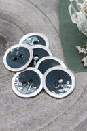 Boutons ronds à deux trous, finition émaillée vert forêt sombre cerclée d'un liseré blanc nacré, présentés en tas sur une pierre grise, avec une feuille verte et une petite coupelle en verre facetté en arrière-plan.