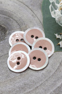 Two-hole round buttons, sandy rosy-beige enamel finish rimmed with a pearly white trim, piled on a green leaf, with a small faceted glass dish and white flower petals in the background.