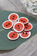 Two-hole round buttons, vivid coral orange enamel finish rimmed with a pearly white trim, piled on a green leaf, with a small faceted glass dish and white flower petals in the background.