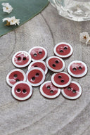 Two-hole round buttons, terracotta red enamel finish rimmed with a pearly white trim, piled on grey stone, with a green leaf and a small faceted glass dish in the background.