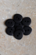 Sept petits boutons ronds mats à deux trous, coloris noir mat, présentés en tas dans une coupelle en pierre ovale beige marbrée.