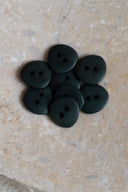 Sept petits boutons ronds mats à deux trous, coloris vert forêt très foncé, présentés en tas dans une coupelle en pierre ovale beige marbrée.
