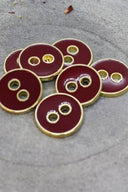 Cinq boutons ronds à deux trous, finition émaillée brillante bordeaux amarante, cerclés d'un liseré métallique doré, posés sur une pierre grise avec des pétales de fleurs séchées éparpillés en arrière-plan.