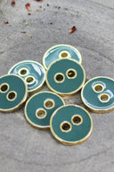 Five two-hole round buttons, glossy cactus green enamel finish, rimmed with a metallic gold trim, set on grey stone with scattered dried flower petals in the background.