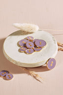 Two-hole round buttons, glossy mauve purple enamel finish rimmed with a metallic gold trim, piled in a small white ceramic dish on a pink background with a dried lagurus spike.