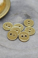 Sept petits boutons ronds à deux trous, finition pailletée dorée sur fond jaune moutarde doré, cerclés d'un liseré métallique doré, posés sur une pierre grise, avec une coupelle dorée floutée en arrière-plan.