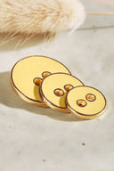Three two-hole round buttons in graduated sizes, glossy golden lemon yellow enamel finish, rimmed with a metallic gold trim, set on light-colored stone with a sprig of dried lagurus grass in the background.