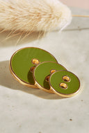 Three two-hole round buttons in graduated sizes, glossy olive matcha green enamel finish, rimmed with a metallic gold trim, set on light-colored stone with a sprig of dried lagurus grass in the background.