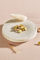 Irregularly shaped, faceted stone-effect buttons in metallic golden lemon yellow, with a gold metal shank, piled in a small white ceramic dish on a pink background with dried lagurus spikes.