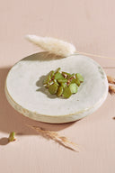 Irregularly shaped, faceted stone-effect buttons in olive matcha green, with a gold metal shank, piled in a small white ceramic dish on a pink background with dried lagurus spikes.