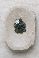 Boutons ronds bombés à queue, sans trous apparents, finition nacrée vert cèdre grisé, présentés en tas de tailles variées dans une coupelle en pierre ovale beige marbrée.