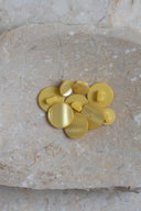 Boutons ronds bombés à queue, sans trous apparents, finition nacrée jaune citron doré, présentés en tas sur une petite coupelle en céramique blanche, sur fond rose avec un épi de lagurus séché.