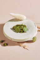 Boutons ronds bombés à queue, sans trous apparents, finition nacrée vert matcha olive, présentés en tas sur une petite coupelle en céramique blanche, sur fond rose avec un épi de lagurus séché.