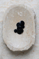 Boutons ronds bombés à queue, sans trous apparents, finition nacrée noir intense (night), présentés en tas de tailles variées dans une coupelle en pierre ovale beige marbrée.