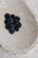 Une douzaine de petits boutons ronds brillants à deux trous, coloris noir anthracite profond, présentés en tas dans une coupelle en pierre beige marbrée.