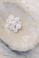 Une douzaine de petits boutons ronds brillants à deux trous, coloris blanc cassé nacré, présentés en tas dans une coupelle en pierre beige marbrée.