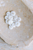 Une douzaine de petits boutons ronds brillants à deux trous, coloris blanc optique pur, présentés en tas dans une coupelle en pierre beige marbrée.