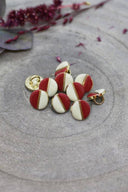 Boutons ronds bicolores à queue métallique dorée, moitié blanc cassé et moitié rouge terracotta, séparés par un fin liseré doré, présentés en tas sur une pierre grise avec un rameau de fleurs séchées bordeaux en arrière-plan.