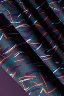 Fuji fabric in anthracite with graphic strokes in orange, sky blue and teal, draped over a white stand near a decorative ornamental onion flower.