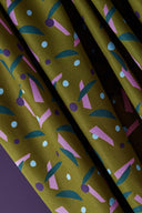 Java fabric in khaki with a graphic confetti print in pink, blue and plum, draped over a white stand near a decorative ornamental onion flower in a green ceramic vase.