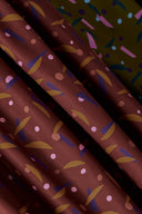 Tissu Java bordeaux à imprimé confettis graphiques rose, moutarde et prune, drapé sur un socle blanc près d'une fleur d'ail décoratif.