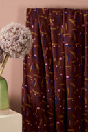 Tissu Java bordeaux drapé sur un socle blanc, avec un vase en céramique verte et une fleur d'ail décoratif en arrière-plan.