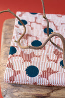 Mirage fabric in off-white with a graphic pattern of fine stripes and polka dots in terracotta and navy, folded on a wooden stand with dried twigs, against a terracotta red background.